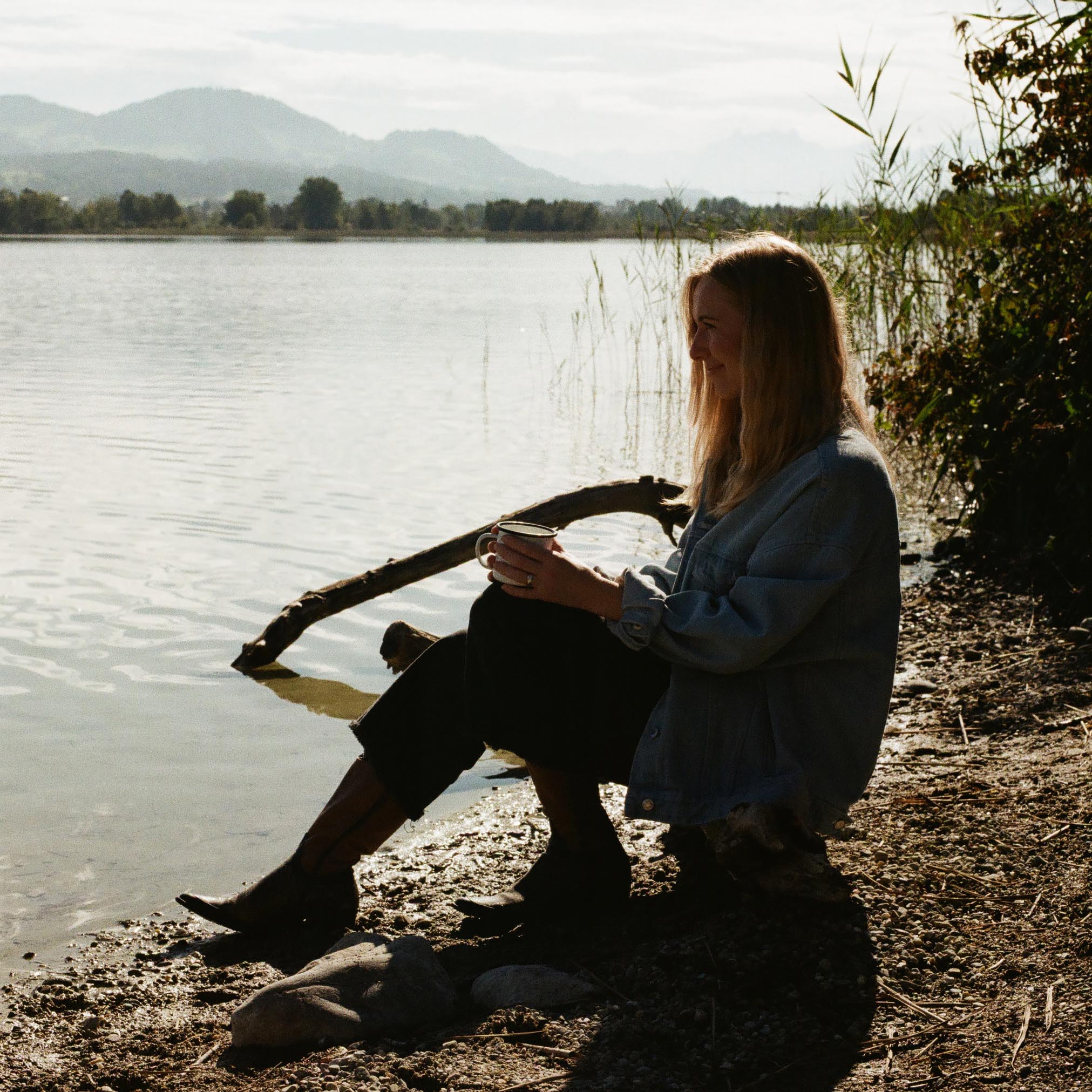 women sitting at the lake shore looking into the distance while holding a coffee cup